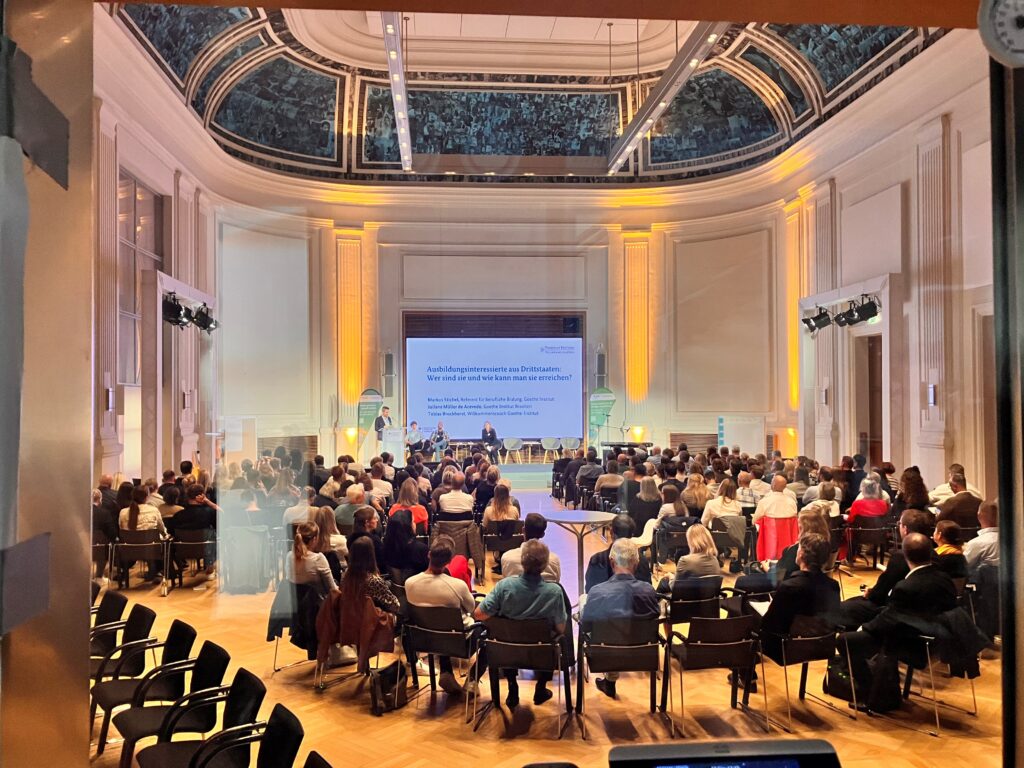 Ein Konferenzsaal mit mehreren Reihen von Teilnehmern, die auf eine Bühne schauen. Auf der Bühne findet eine Podiumsdiskussion statt. Auf der Leinwand im Hintergrund steht der Titel: ‚Ausbildungsinteressierte aus Drittstaaten: Wer sind sie und wie kann man sie erreichen?‘.