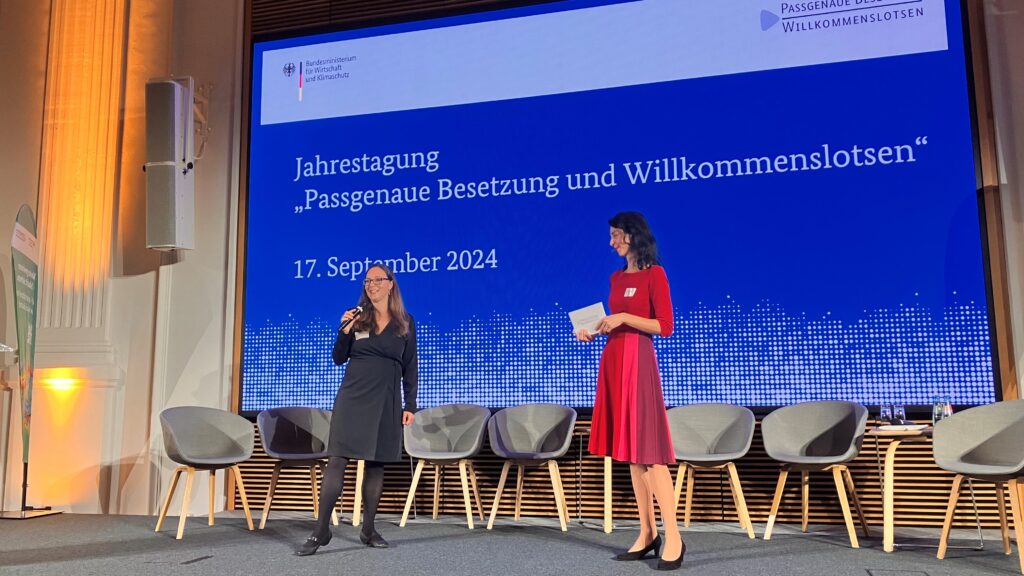 Zwei Frauen stehen auf einer Bühne vor einer Leinwand, auf der der Text ‚Jahrestagung Passgenaue Besetzung und Willkommenslotsen, 17. September 2024‘ angezeigt wird. 